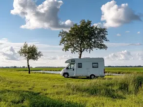 Bauernhof-Romantik: Mit dem Campervan ins Naturabenteuer - © Oksana, adobe stock Ein weißer Campervan steht inmitten einer lebendigen Landschaft, eingerahmt von zwei Bäumen unter einem Himmel mit flauschigen Wolken – das Bild strahlt ein Gefühl von Freiheit aus.