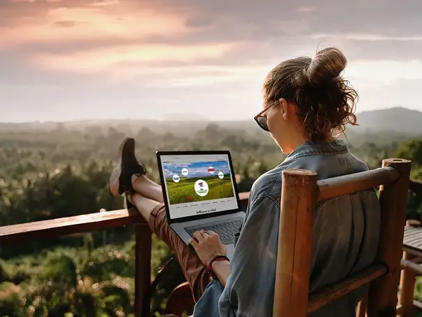 Ical Schnittstelle Frau auf Terasse mit Blick über Wald arbeitet an Laptop. Auf dem Bildschirm sieht man symbolisch die Anbindemöglichkeiten mit Ical.