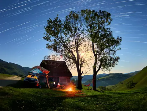 Camping auf dem Bauernhof - © thecriss, adobe stock Ein Auto mit Dachzelt steht zwischen zwei großen Bäumen, daneben ein kleines, gemütlich brennendes Lagerfeuer. Über der Szene spannt sich ein klarer Nachthimmel voller funkelnder Sterne.