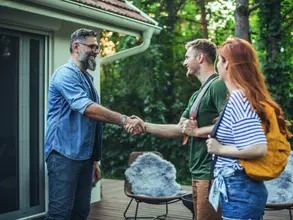 1017057100 Young couple renting house. House owner shaking hands with couple who rent his house. Couple is on vacation and they are wearing backpacks.