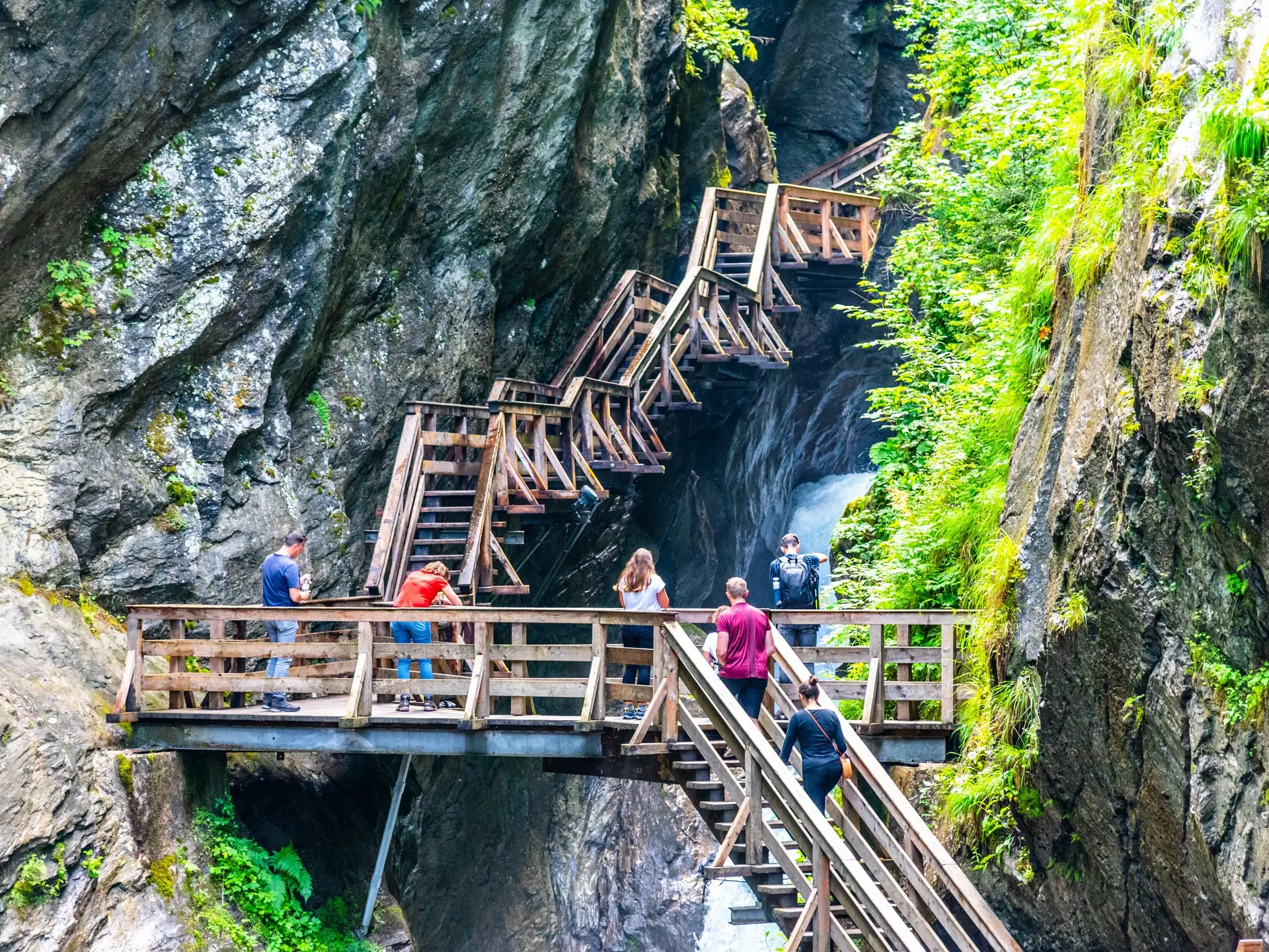 Auf steilen Holztreppen in einer felsigen Klamm stehen mehrere Menschen und schauen nach unten.