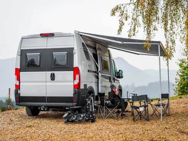 Herbstzauber auf dem Bauernhof – Mit dem Camper in die Hügel - © _jure, adobe stock Ein weißer Camper mit Markise und Campingstühlen lädt zum Verweilen ein – goldene Herbstlandschaft und hügelige Kulisse im Hintergrund.