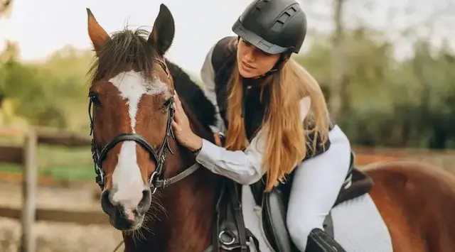 Ferien auf dem Reiterhof - © © sergo321 - AdobeStock Frau mit langen Haaren sitzt in Reiteranziehsachen auf braunem Pferd und streichelt es am Hals.