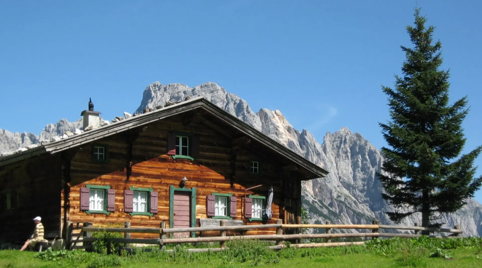 Rustikale Holzhütte mit goßer Fichte nebendrann und wilden Felsformationen dahinter.