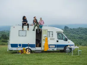 Freiheit erleben – Camping auf dem Bauernhof im Campervan - © PintoArt, adobe stock Auf einer grünen Wiese, vor dem Hintergrund eines Waldes, sitzen drei junge Menschen entspannt auf dem Dach eines weißen Campervans – ein Moment voller Freiheit und Naturverbundenheit.