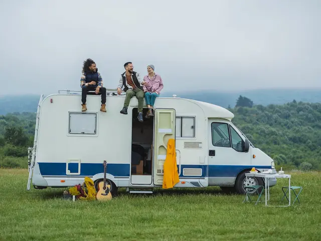Freiheit erleben – Camping auf dem Bauernhof im Campervan - © PintoArt, adobe stock Auf einer grünen Wiese, vor dem Hintergrund eines Waldes, sitzen drei junge Menschen entspannt auf dem Dach eines weißen Campervans – ein Moment voller Freiheit und Naturverbundenheit.