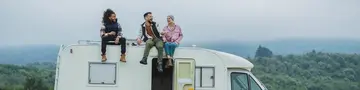 Freiheit erleben – Camping auf dem Bauernhof im Campervan - © PintoArt, adobe stock Auf einer grünen Wiese, vor dem Hintergrund eines Waldes, sitzen drei junge Menschen entspannt auf dem Dach eines weißen Campervans – ein Moment voller Freiheit und Naturverbundenheit.