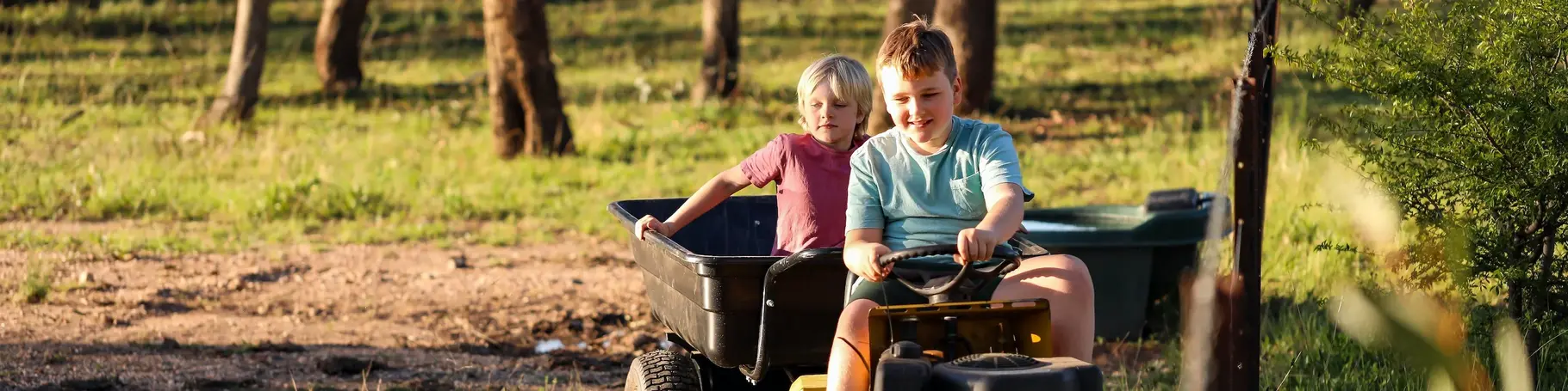 Zwei Jungs die glücklich auf einem Tretttraktor fahren. Der zweite Junge lässt sich in einem Anhänger durch die Natur ziehen.