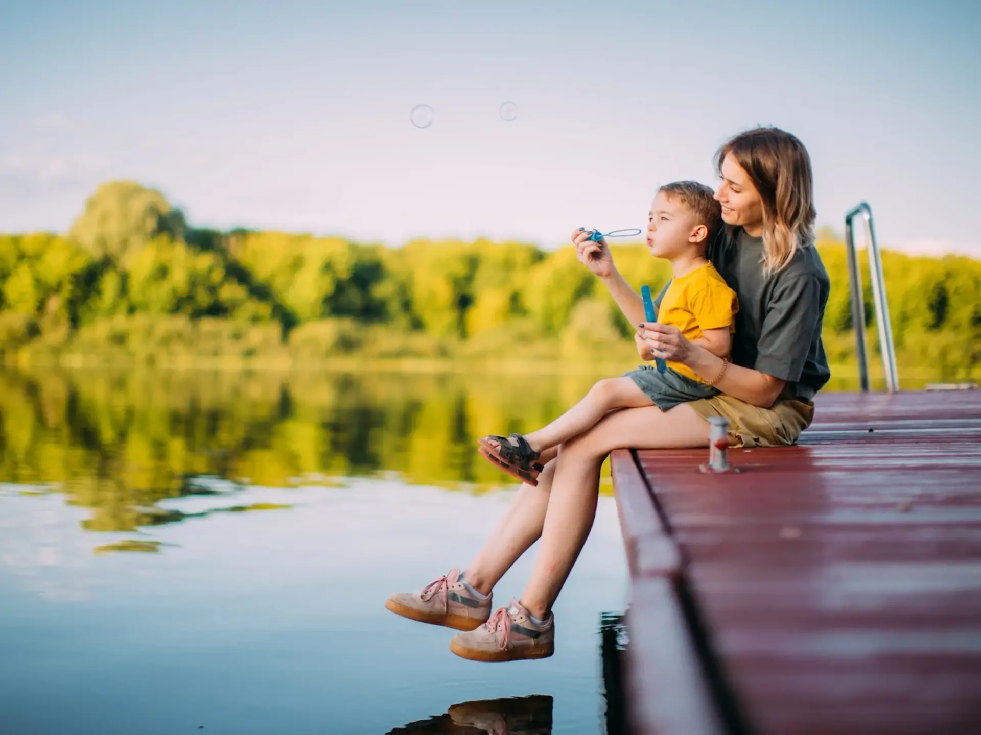 Unbeschwerte Familienzeit am Ferienhaus in der Natur Mutter und kleiner Junge sitzen auf einem Holzsteg in ländlicher Natur und lassen Seifenblasen steigen – idyllische Szene im Sommerurlaub beim Ferienhaus auf dem Land.