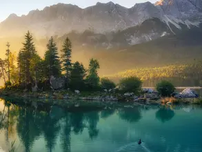 Idyllischer See bei Sonnenaufgang - © Smileus – stock.adobe.com Idyllischer See bei Sonnenaufgang, ein malerisches Panorama mit majestätischen Bergen und dem goldenen Sonnenlicht, das hinter den Bäumen hereinscheint