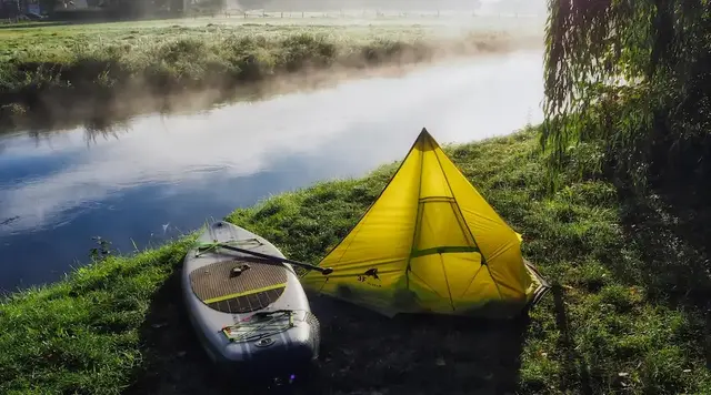 Naturcamping am Fluss – Aktivurlaub auf dem Land - © Svenja, adobe stock Ein gelbes Zelt und ein Paddleboard stehen am nebligen Flussufer während eines naturnahen Urlaubs auf dem Land.