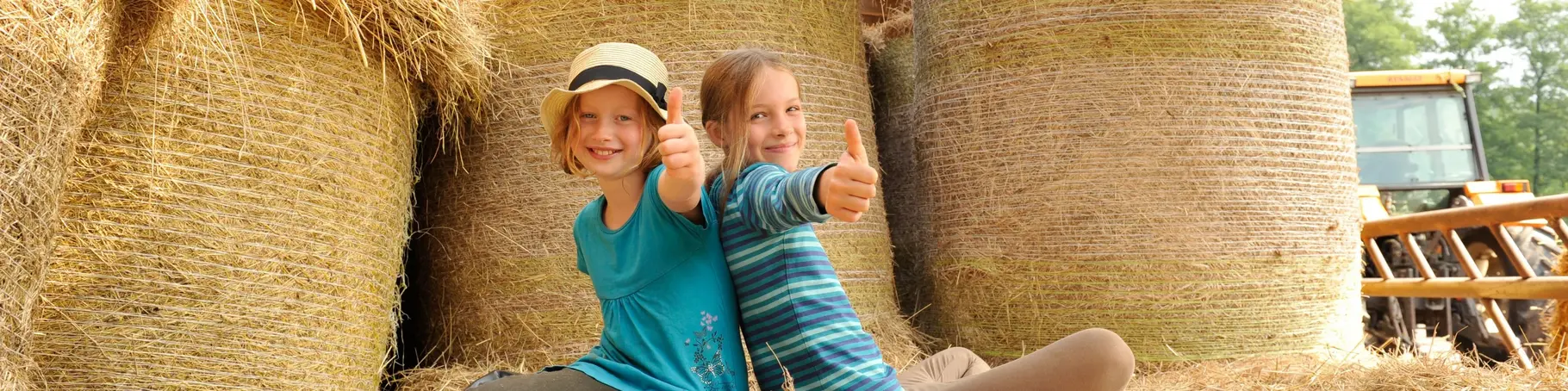 Zwei glückliche Mädchen sitzen auf Heuballen in der Scheune und halten ihre Daumen nach oben