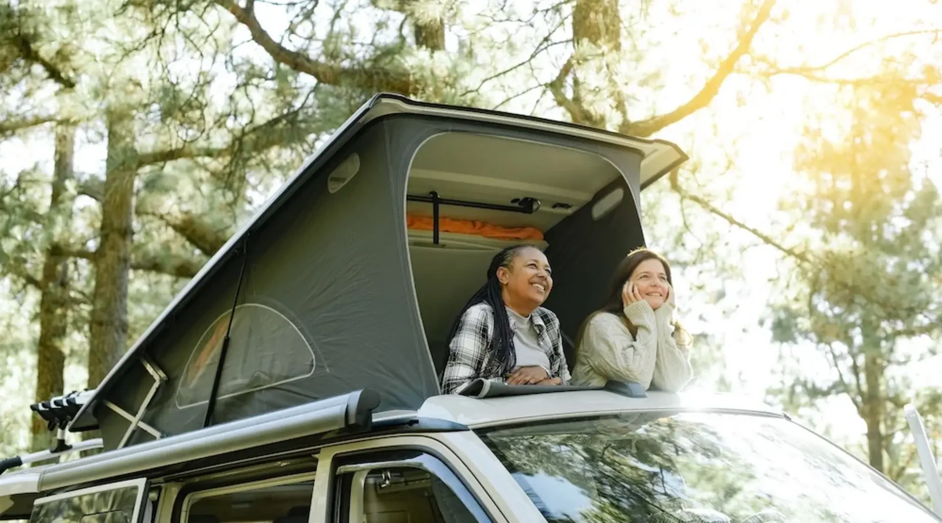 Zwei Frauen schauen aus dem geöffneten Klappdach einen Campingbuses in die von Bäumen bewachsene Umgebung.