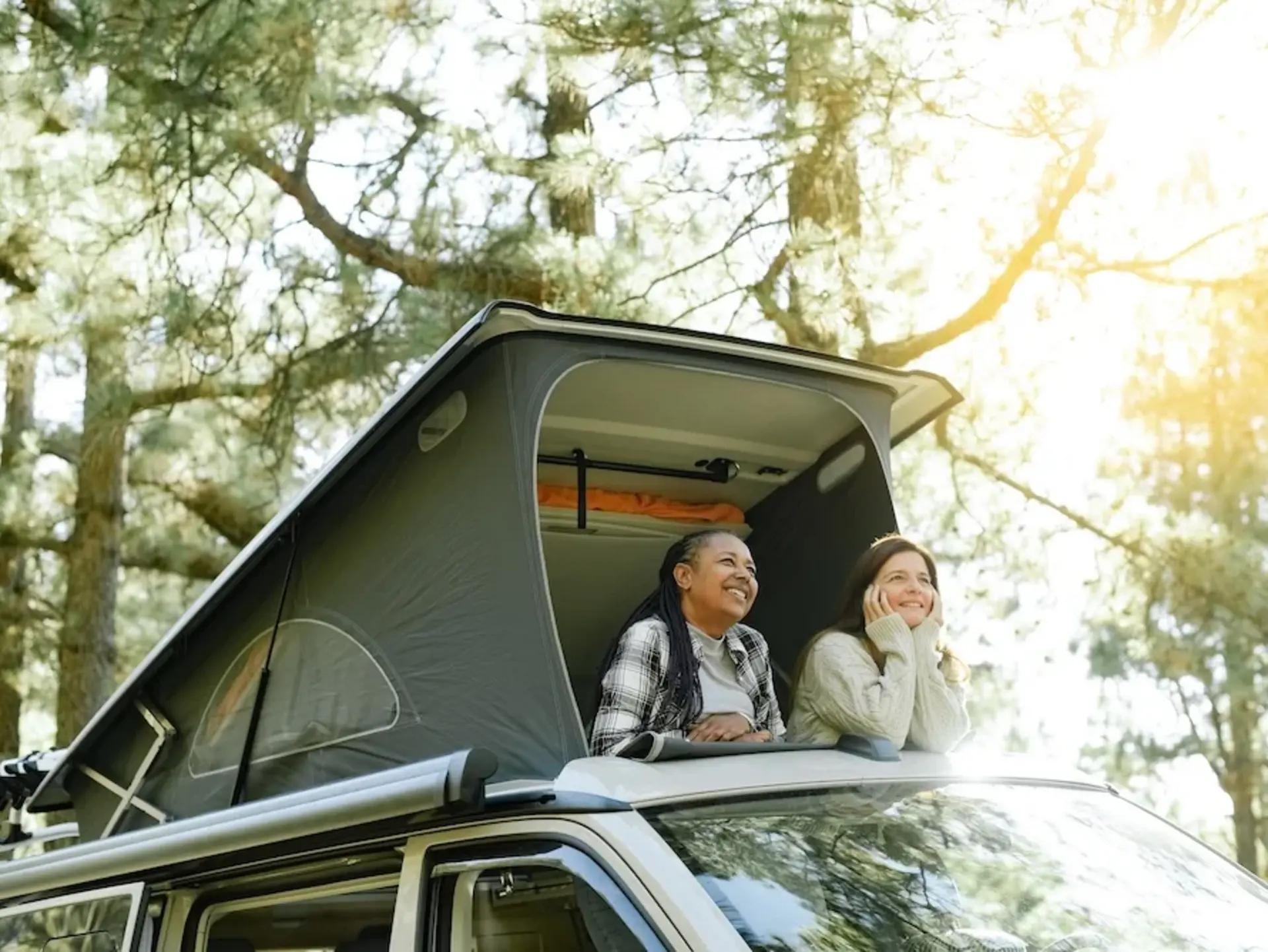 Wunderschöne Natur erleben eim Camping auf dem Bauernhof - © Gabriel Trujillo, adobe stock Zwei Frauen schauen aus dem geöffneten Klappdach einen Campingbuses in die von Bäumen bewachsene Umgebung.
