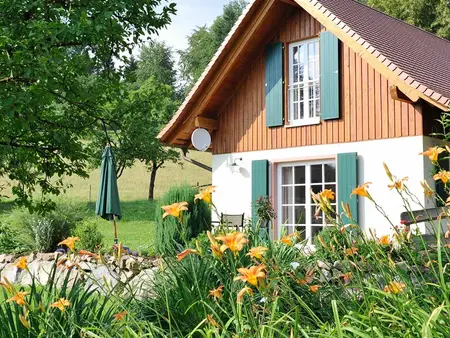 Kleines Haus mit grünen Fensterläden und Holzschirm umgeben von Natur und gelben Lilien