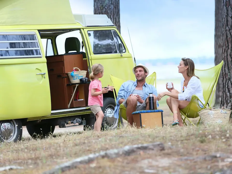 Wohnmobilstellplätze am Bauernhof - © goodluz, adobe stock Ein grüner, nostalgischer Campingbus steht auf sandigem Boden unter schattigen Bäumen. Davor sitzt eine Familie entspannt in Campingstühlen und genießt die Natur.