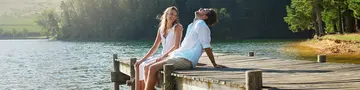 Young couple, lake and jetty with laughing, happiness and bonding with love in nature for holiday. Man, woman and comic joke on bridge by water to relax with conversation, care and vacation in summer